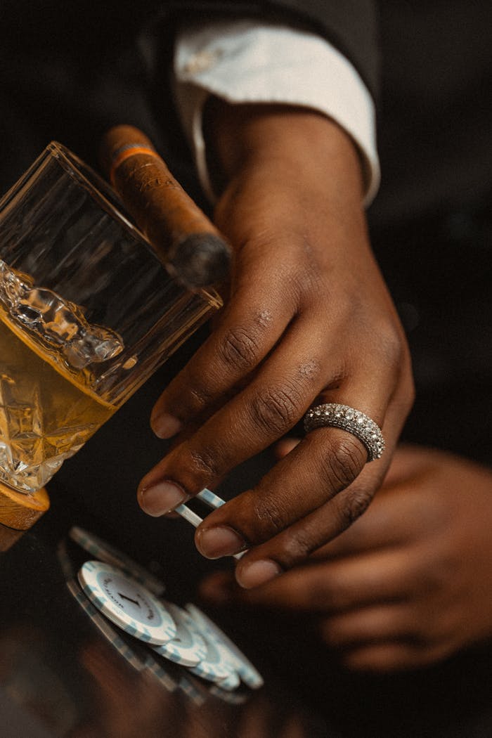 A close-up of a man's hand with a whisky glass, cigar, and poker chips, exuding elegance.